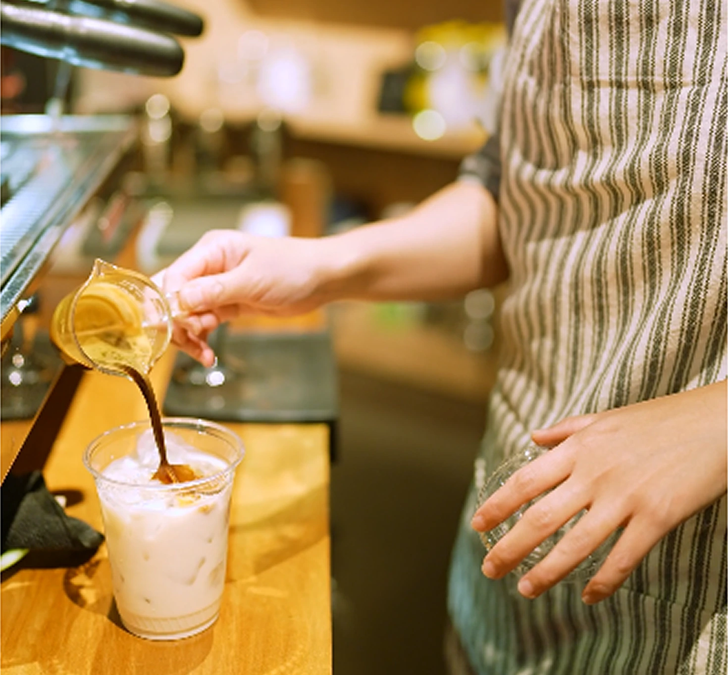 Barista in striped apron pouring espresso into a plastic cup of iced milk to make iced coffee.